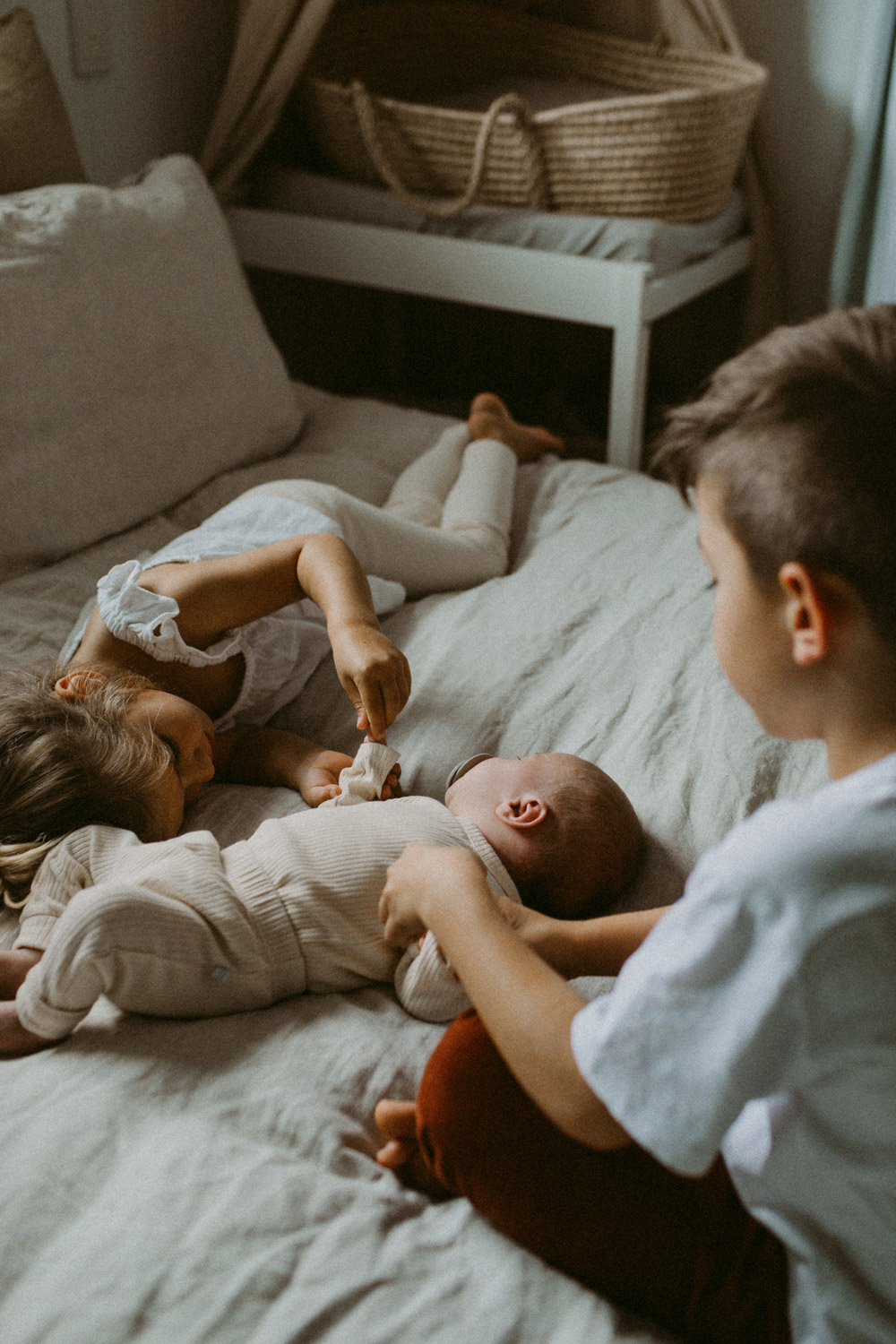 candid photo of siblings holding newborn babies hands on bed at home